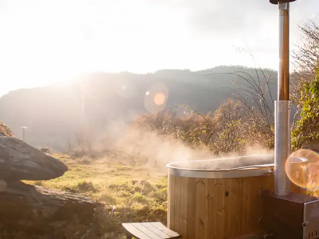 Ein dampfender Holzzuber steht draußen in einer sonnigen, hügeligen Landschaft.