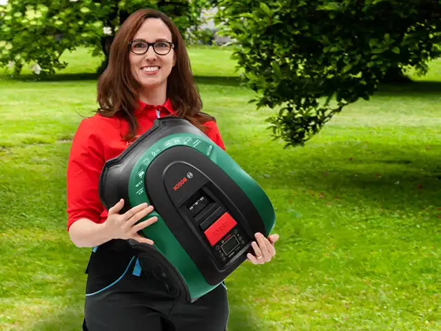 Eine Frau hält einen automatischen Rasenmäher von Bosch in einem sonnigen, grünen Garten.