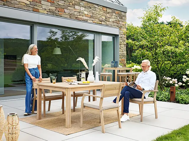 Ein Tisch steht auf einer Terrasse, umgeben von Pflanzen; zwei Personen interagieren entspannend.