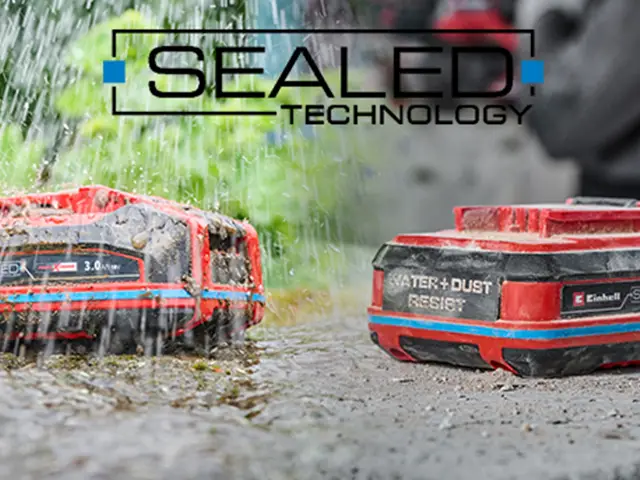 Zwei Einhell-Akkus widerstehen starkem Regen auf nasser Oberfläche, umgeben von grüner Vegetation.