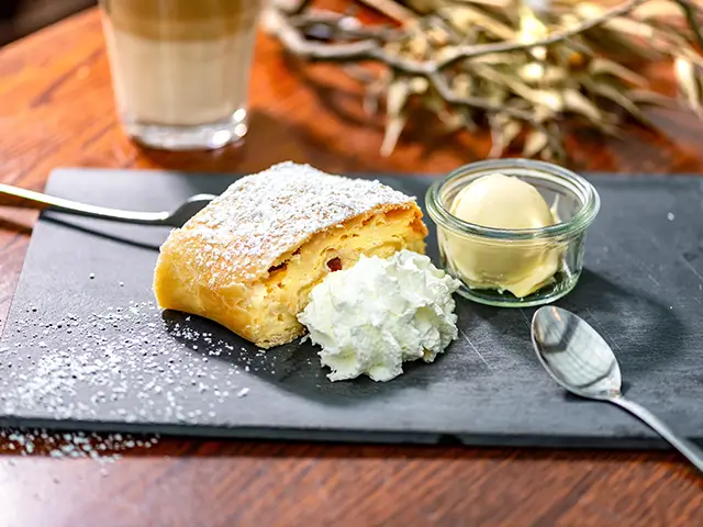 Ein Stück Apfelstrudel mit Sahne und Vanilleeis auf einer Schiefertafel und einem Latte Macchiato im Hintergrund.