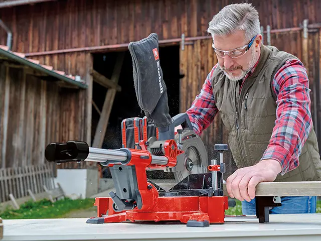 Ein Mann schneidet Holz auf einer roten Einhell Gehrungssäge in einem Hof mit Holzgebäude.