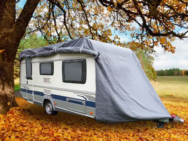 Ein Wohnwagen ist von einer Abdeckung bedeckt und steht unter herbstlichen Bäumen auf Laub.