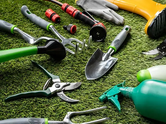 Gartengeräte liegen verstreut auf einem grünen Rasen, darunter eine Schaufel, eine Schere und eine Gießkanne.