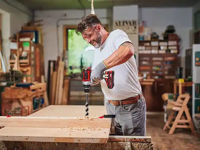 Ein Mann bohrt mit einer Einhell Bohrmaschine Löcher in ein Holz, mitten in einer Werkstatt mit Regalen und Werkzeugen.