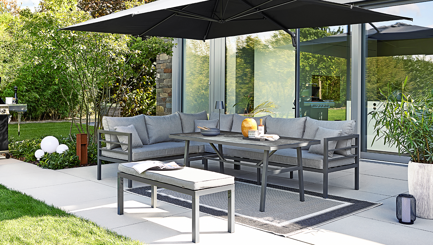 Gartentisch und Sofas stehen unter einem großen Sonnenschirm auf einer modernen Terrasse.