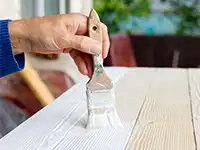 Eine Hand streicht mit einem Pinsel eine weiße Farbe auf eine Holzoberfläche im Freien.