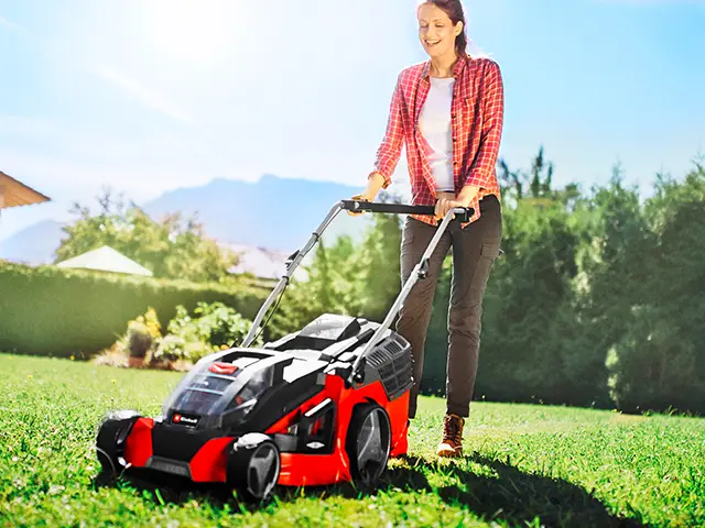 Eine Frau schiebt einen roten Einhell Rasenmäher im sonnigen Garten.