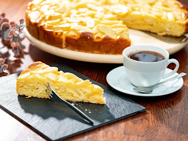 Ein Stück Apfelkuchen liegt neben einer Tasse Kaffee auf einem Holztisch.