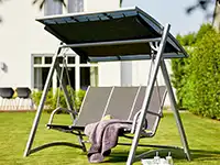 Eine Gartenschaukel mit Dach steht auf einem Rasen vor einem Wohnhaus.