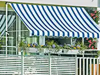 Eine blau-weiß gestreifte Markise überdeckt einen Balkon mit Blumen in Pflanzkästen.