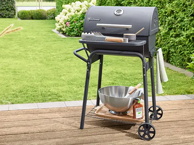 Ein Rothmann Holzkohlegrill steht auf der Terrasse, umgeben von einem grünem Garten und Pflanzen.
