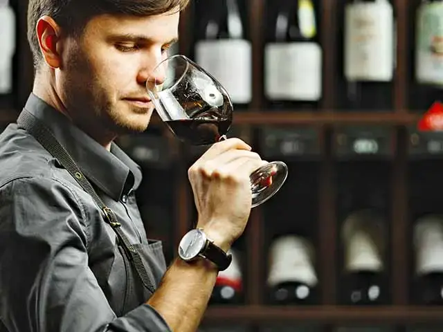 Ein Mann riecht an einem Glas Rotwein, umgeben von Weinflaschen im Hintergrund.