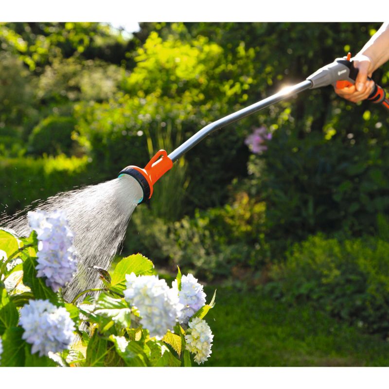 Eine Person bewässert mit einem Gardena Gießstab weiße Blumen in einem sonnigen Gartenumfeld.