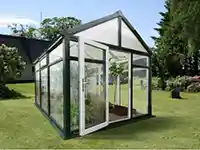 Ein Gewächshaus steht auf einer Wiese, umgeben von Bäumen und Sträuchern.