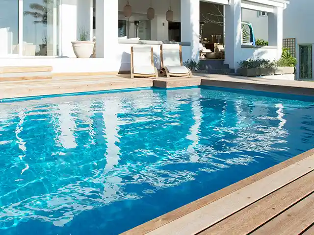 Ein rechteckiger Pool reflektiert Sonnenlicht, umgeben von einer Holzterrasse und einem weißen Gebäude.