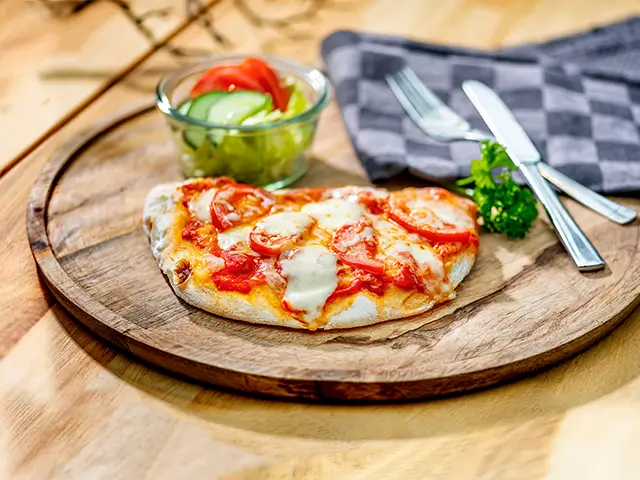Eine Pinsa liegt auf einem Holzbrett mit Salat, Besteck und Serviette im Hintergrund.