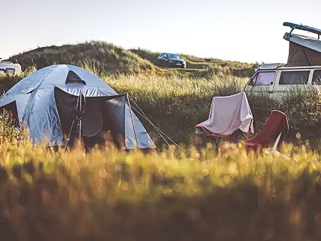 Ein Zelt steht neben Campingstühlen auf einer Wiese mit Autos im Hintergrund.