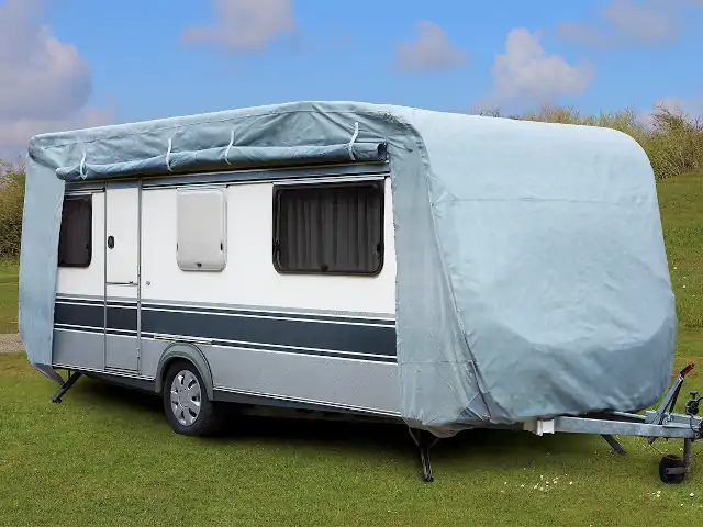 Ein Wohnwagen steht auf einer grünen Wiese, mit einer grauen Plane bedeckt.