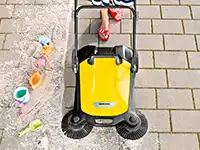 Eine gelbe Kehrmaschine beseitigt Sand von einem gepflasterten Gehweg.