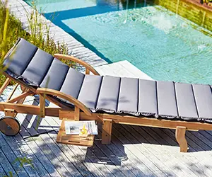 Eine Sonnenliege mit grauem Polster steht neben einem Pool auf einem Holzdeck.
