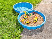 Ein Sandkasten in Muschelform mit Spielzeugtraktoren steht auf Gras neben einem Gehweg.