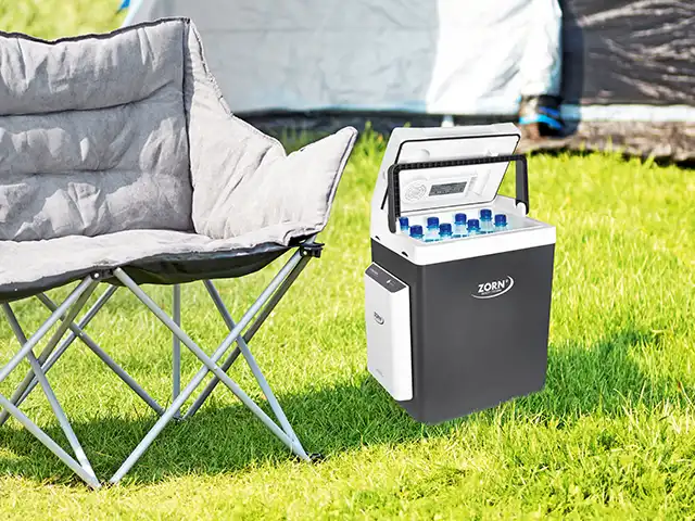 Ein Campingstuhl steht neben einer geöffneten Kühlbox mit Flaschen auf grünem Gras.