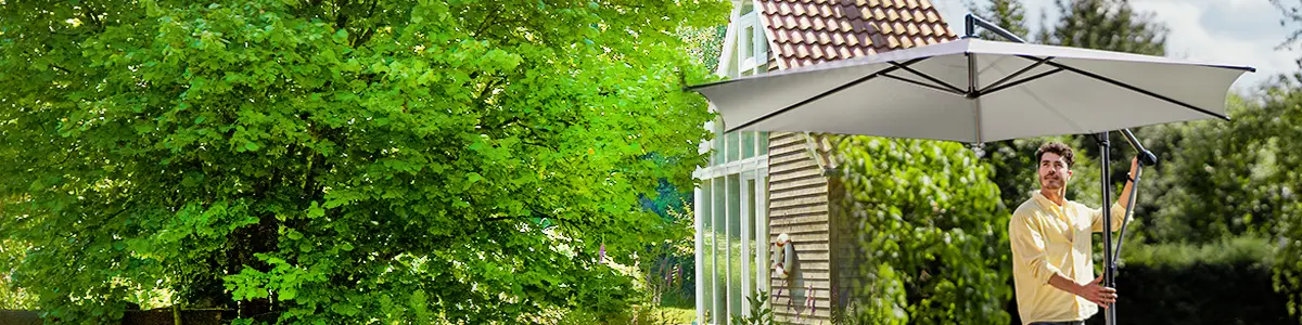 Ein Mann öffnet einen großen Sonnenschirm im Garten neben einem Glashaus.
