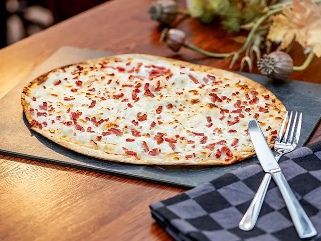 Ein Flammkuchen mit Speck liegt auf einem Tisch, daneben Besteck und eine Serviette.