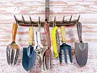 Verschiedene Gartengeräte hängen an einer Harke vor einer verwitterten Holzwand.