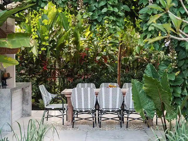 Ein Esstisch mit Stühlen steht unter üppigen, grünen Pflanzen im Freien.