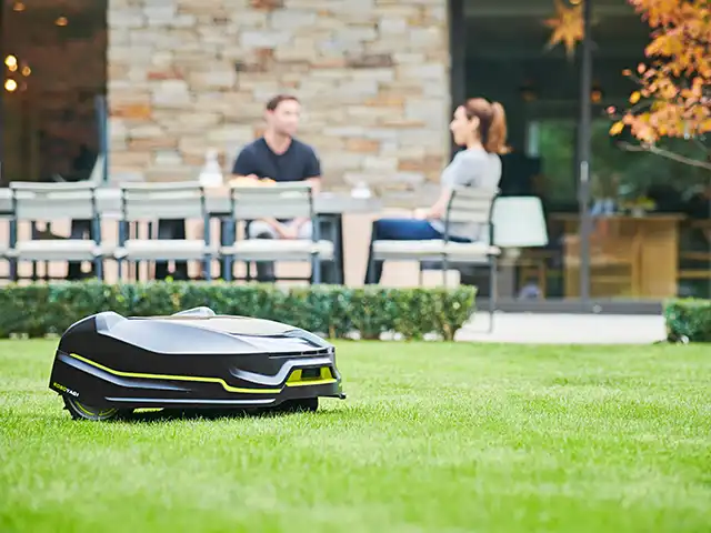 Ein Rasenmähroboter mäht Gras im Garten, während zwei Personen am Tisch auf der Terrasse sitzen.