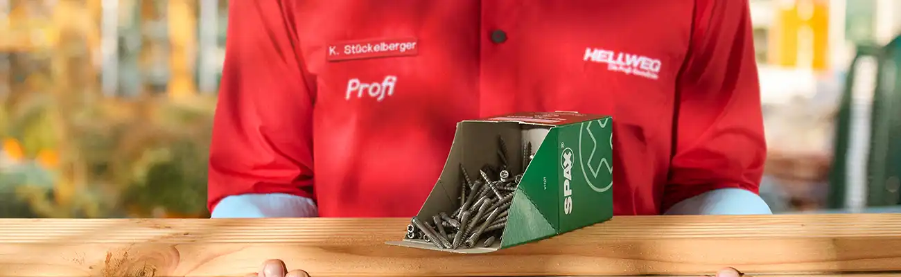 Ein Hellweg Mitarbeiter hält ein Holzstück, auf dem sich eine Kiste Schrauben befindet.