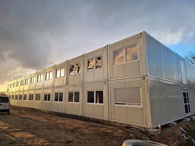 Ein zweistöckiges Containergebäude steht auf einer Baustelle bei bewölktem Himmel.