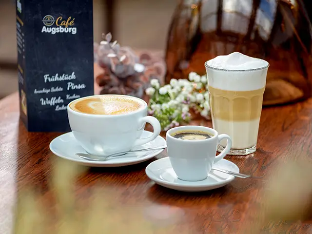 Ein Espresso, Cappuccino und Caffè Latte stehen auf einem Holztisch im Café Augsburg.