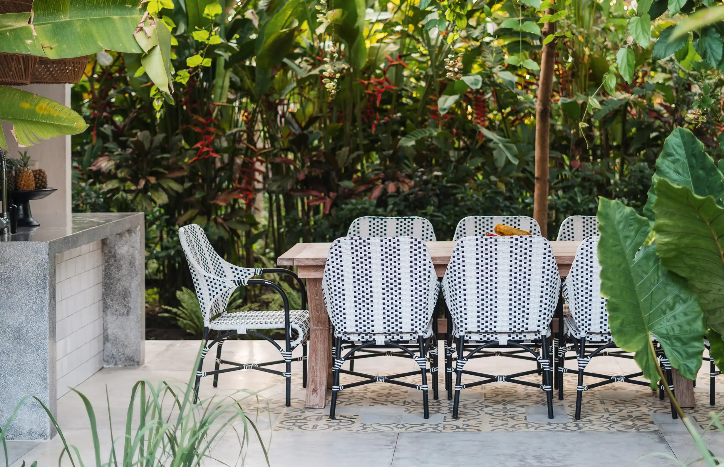 Ein Tisch mit geflochtenen Stühlen steht im Freien, umgeben von üppigen, grünen Pflanzen.