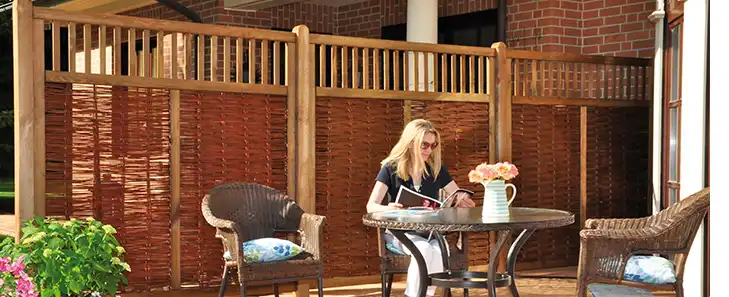Eine Frau sitzt lesend an einem Tisch auf einer Terrasse mit Holzsichtschutz.