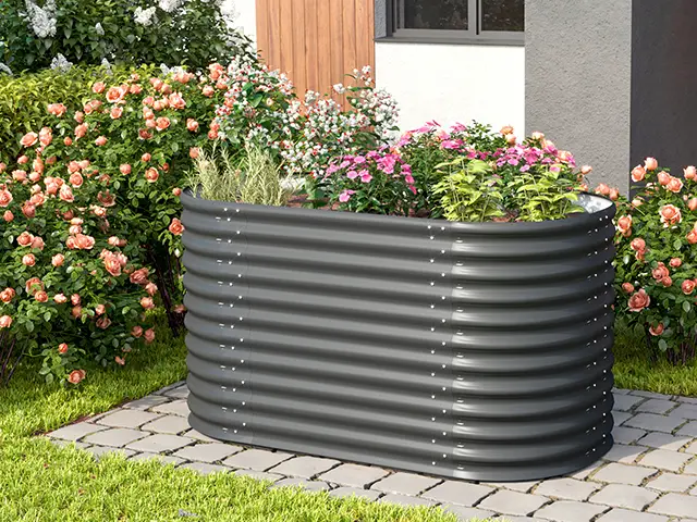 Ein graues Hochbeet mit blühenden Pflanzen steht in einem gepflasterten Gartenabschnitt, umgeben von Blumen.