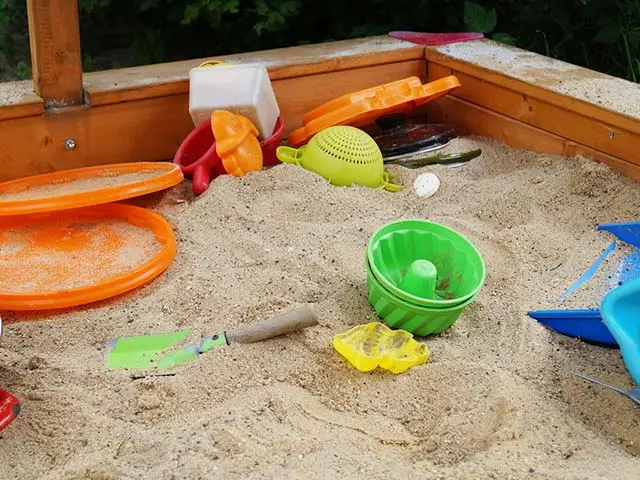 Bunte Spielzeuge liegen verstreut in einem Sandkasten, umgeben von grüner Vegetation.