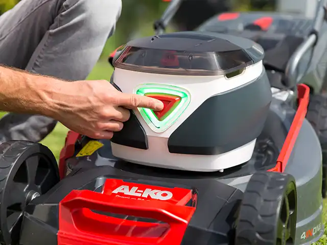 Eine Person drückt eine Taste auf einem AL-KO Rasenmäher in einem Gartenumfeld.