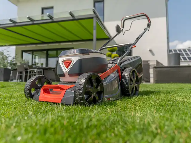 Ein Rasenmäher steht auf grünem Rasen vor einem modernen Haus mit Terrasse.