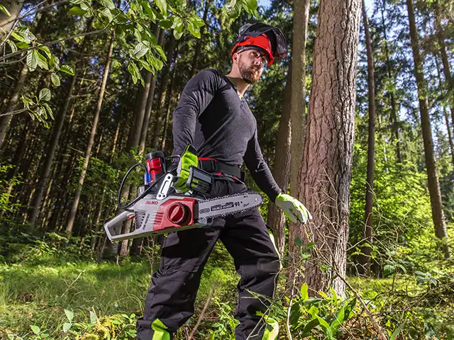 Ein Mann mit einer AL-KO Motorsäge arbeitet mit Schutzkleidung im Wald neben einem Baum.