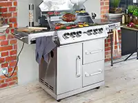 Ein Edelstahlgrill brät Fleisch und Gemüse auf einer Terrasse mit Ziegelwänden.