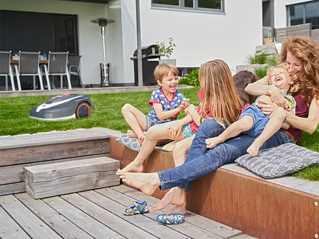 Eine Frau spielt mit Kindern auf einer Terrasse neben einem AL-KO Mähroboter im Garten.