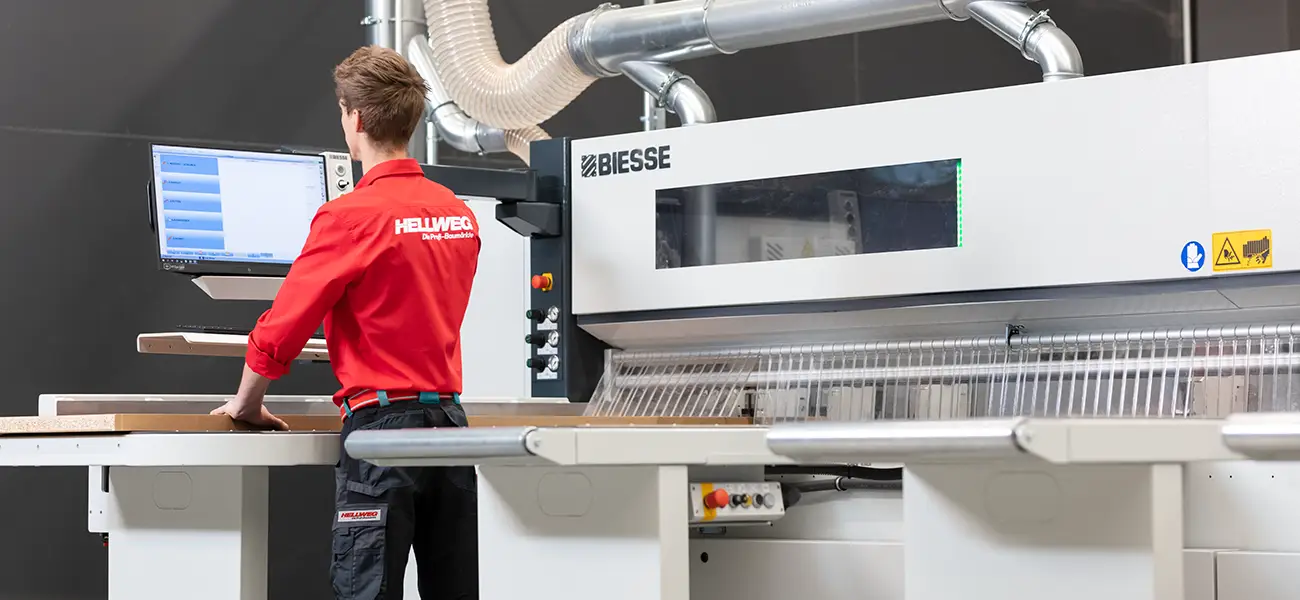 Ein Hellweg Mitarbeiter bedient eine Biesse-Maschine in einer Werkstatt mit Holzplatten.