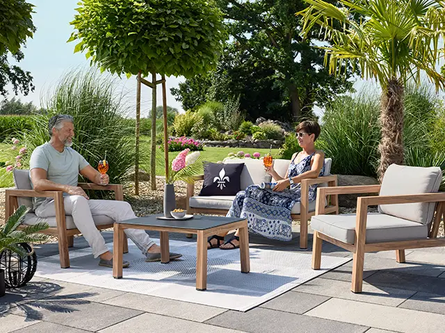 Zwei Menschen sitzen auf Gartenmöbeln und trinken Cocktails in einem sonnigen, grünen Garten.