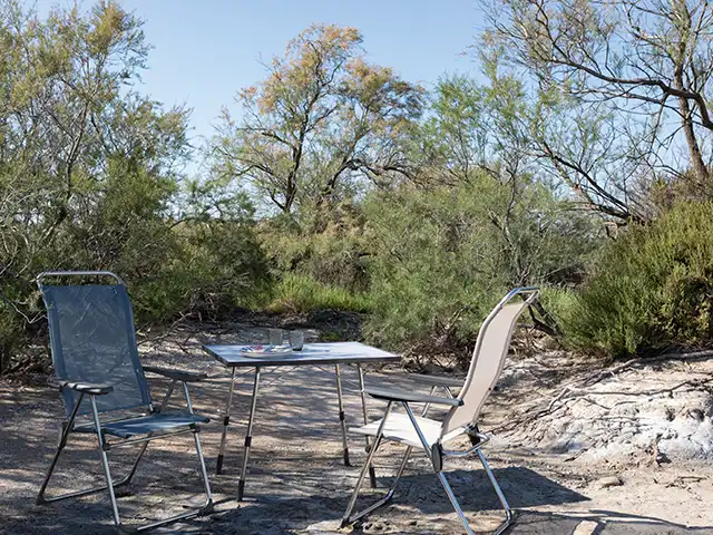 Zwei Stühle stehen um einen Tisch im Freien, umgeben von Büschen und Bäumen.