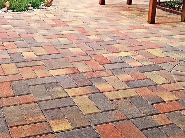 Mehrfarbige Pflastersteine liegen gleichmäßig verlegt in einem Außenbereich neben einem bepflanzten Bereich.