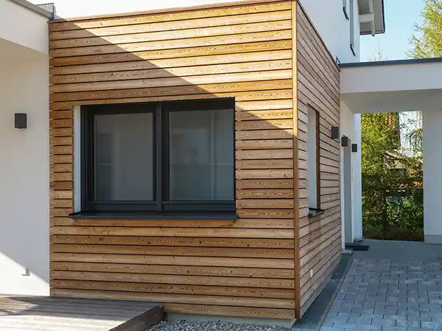 Ein Gebäudeabschnitt mit Holzverkleidung und Fenster steht neben einer gepflasterten Einfahrt im Freien.
