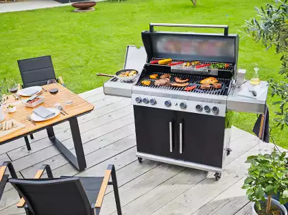 Ein Grill brät Fleisch und Gemüse auf einer Holzterrasse mit Tisch und Stühlen.